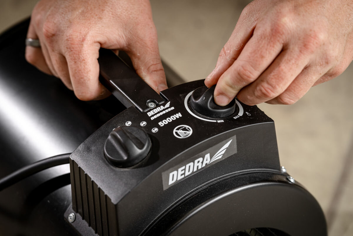 Nagrzewnica elektryczna DEDRA DED9932B, termostat, przełącznik, kąt, precyzja, Ręce ustawiają pokrętło mocy i temperatury na panelu DED9932B, widoczne oznaczenie 5000W.