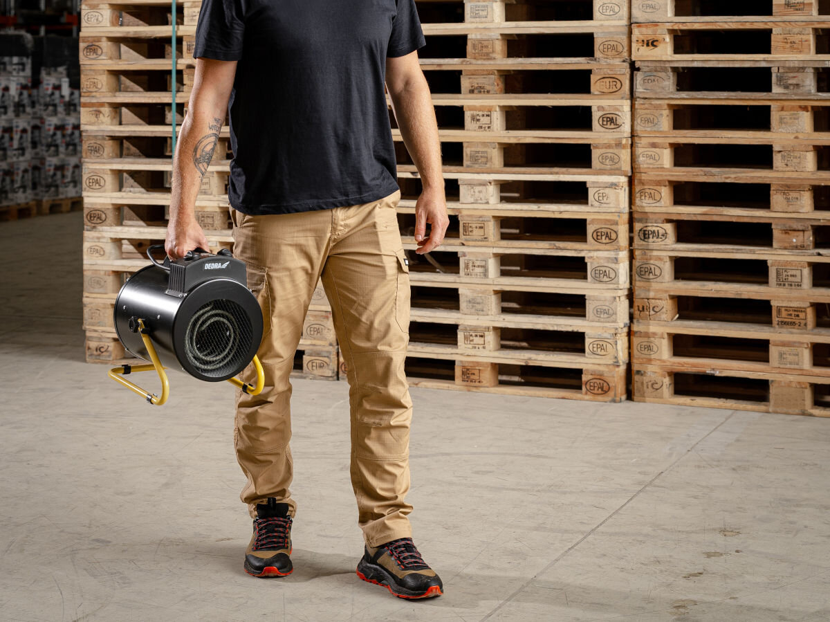 Nagrzewnica elektryczna DEDRA DED9932B, dodatkowy, wentylacja, brud, komfort, Mężczyzna niesie dużą nagrzewnicę DEDRA DED9932B w hali z paletami, widok od przodu.