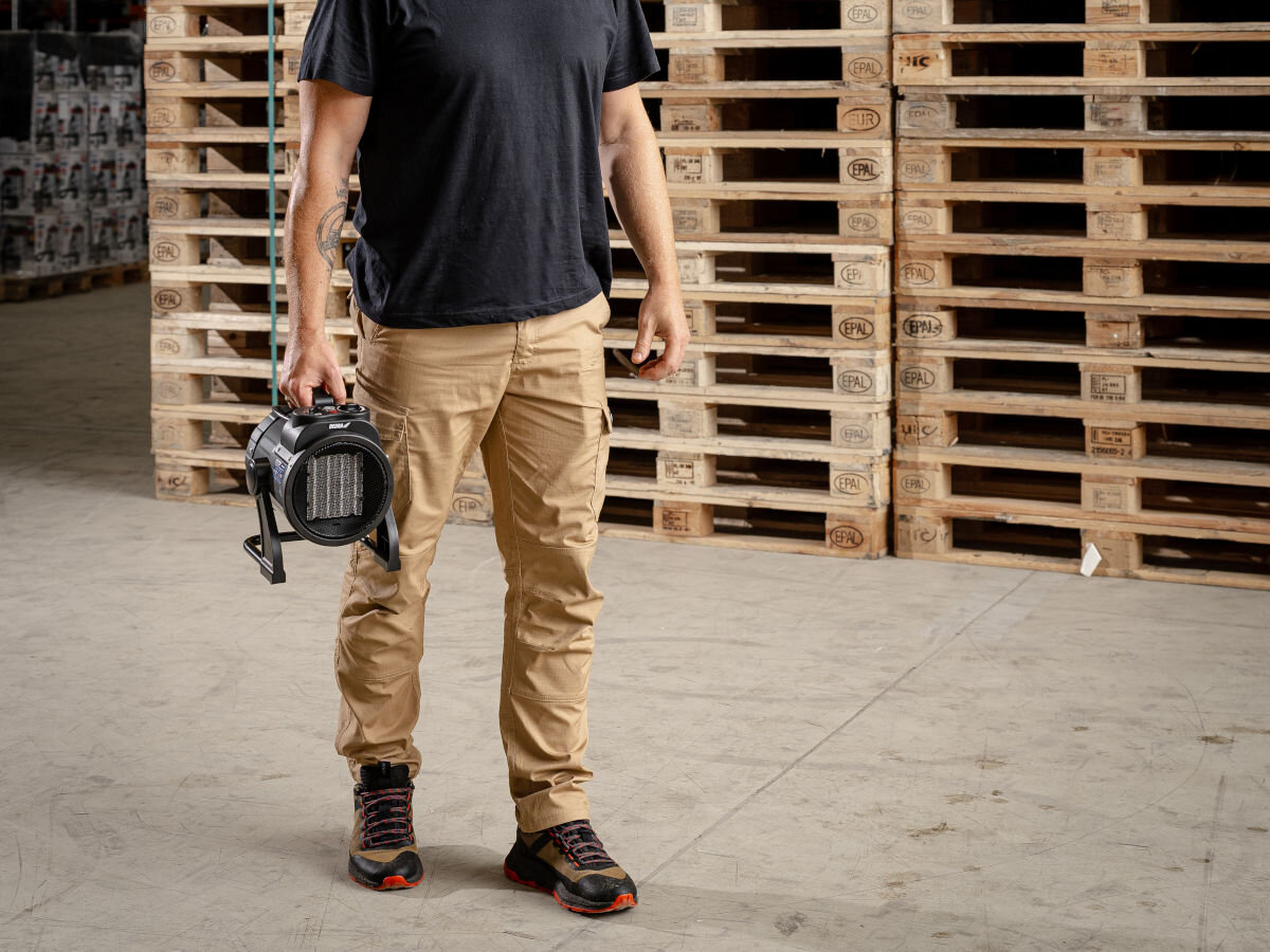 Nagrzewnica elektryczna DEDRA DED9930C2, lekkość, uchwyt, wymiar, ergonomia, Mężczyzna niesie nagrzewnicę Dedra DED9930C2 w hali magazynowej przy paletach, stabilna podłoga.
