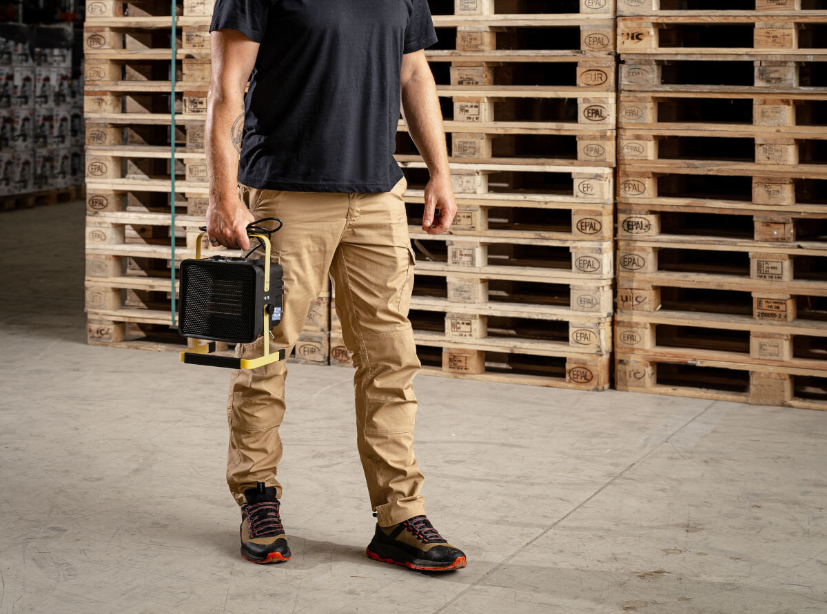 Nagrzewnica elektryczna DEDRA DED9930C3, lekkość, uchwyt, cichość, stabilność, Mężczyzna niesie nagrzewnicę ręką za uchwyt, przemysłowy magazyn, palety w tle.