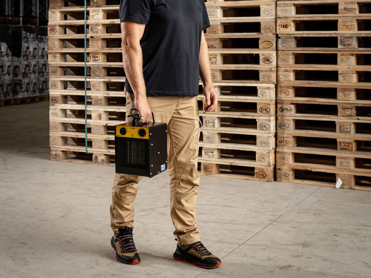 Nagrzewnica elektryczna DEDRA DED9931C1, lekkość, uchwyt, stabilność, cicho, Mężczyzna trzyma w ręce nagrzewnicę Dedra DED9931C1 w hali magazynowej, palety w tle.