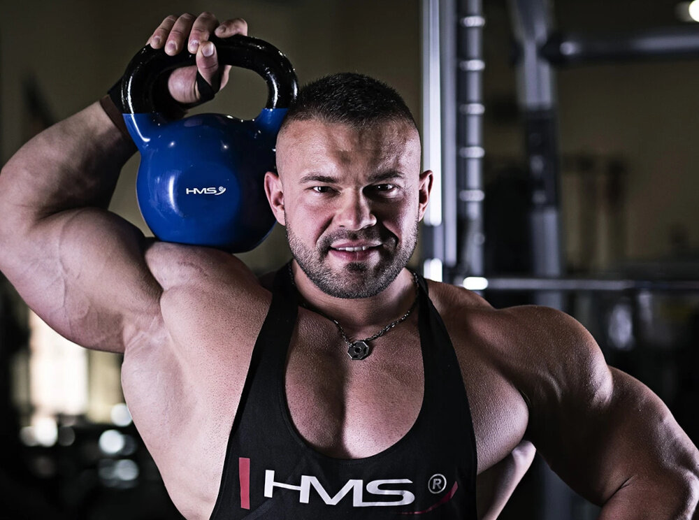 Kettlebell HMS KNV10 (10 kg) Niebiesko-czarny siłownia umięśniona osoba trzyma kettlebell na ramieniu w tle sprzęty do ćwiczeń trening siłowy trening kondycyjny wszechstronne zastosowanie zdrowie