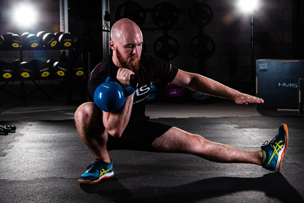 Kettlebell HMS KNV10 (10 kg) Niebiesko-czarny siłownia osoba ćwiczy w półsiadzie z kettlebellem w tle sprzęty do ćwiczeń ćwiczenia wielostawowe z kettlebellem aktywują jednocześnie wiele grup mięśniowych poprawia pracę układu nerwowo-mięśniowego wspiera równowagę oraz koordynację ruchową zmniejszyć ryzyko kontuzji lepszą sprawność