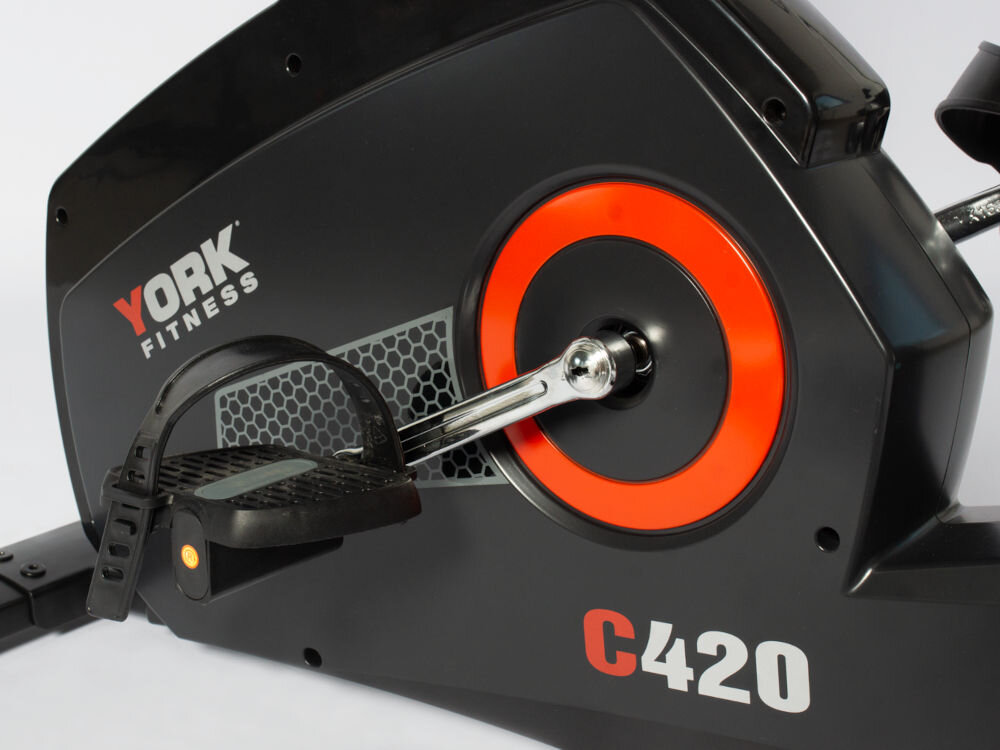 Rower magnetyczny YORK FITNESS C420 z magnetycznym systemem hamowania i 32 poziomami obciążenia elektronicznego Zbłiżenie na pedały z regulowanymi paskami oraz czarny korpus roweru treningowego York C420 z pomarańczowym kołem dekoracyjnym w pobliżu osi.