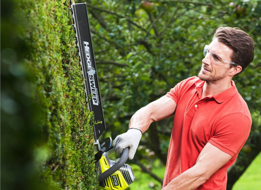 Nożyce do żywopłotu RYOBI RHT6760RL 600mm 0.65kW elektryczne ogród osoba przycina żywopłot w tle drzewa krzewy elektroniczne sprzęgło ochrona przed przypadkowymi blokadami ostrza bezpieczeństwo podczas pracy ogranicza ryzyko nagłych szarpnięć pomaga utrzymać kontrolę nad narzędziem dodatkowa osłona ostrza w kształcie litery T zabezpiecza użytkownika i otoczenie