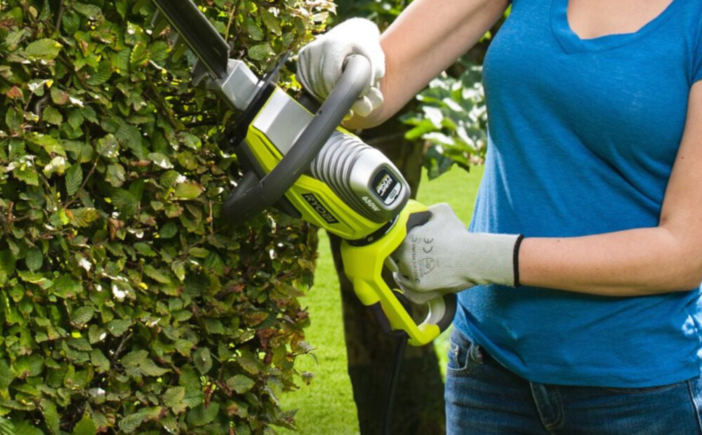 Nożyce do żywopłotu RYOBI RHT6760RL 600mm 0.65kW elektryczne ogród osoba przycina żywopłot w tle drzewa krzewy zbliżenie na uchwyt obrotowy, pięciopozycyjny tylny uchwyt z dużym włącznikiem miękka okładzina dopasowanie pozycji pracy do aktualnego zadania przedni przycisk komfort obsługi przy różnych kątach cięcia