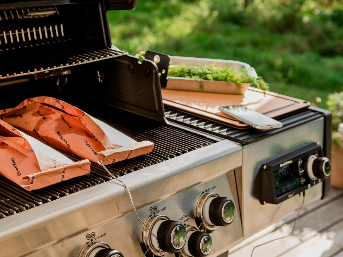 Na zdjęciu widoczny jest grill z otwartą pokrywą, na którym znajdują się dwa papierowe opakowania z napisem \'Broil King\'. Obok grilla na bocznej półce leży deska z ziołami i telefonem.