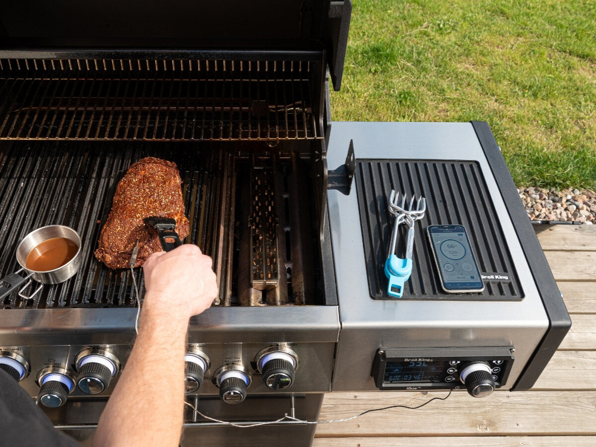Osoba grilluje mięso na dużym grillu gazowym, obok którego znajduje się boczna półka z matą, na której leży telefon i narzędzie kuchenne. Na półce widoczne jest logo \'Broil King\'.
