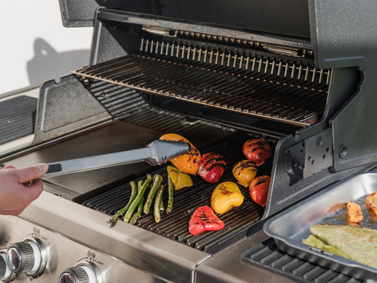 Osoba używa szczypiec do grillowania kolorowych papryk i szparagów na grillu gazowym. Obok znajduje się metalowa taca z jedzeniem.