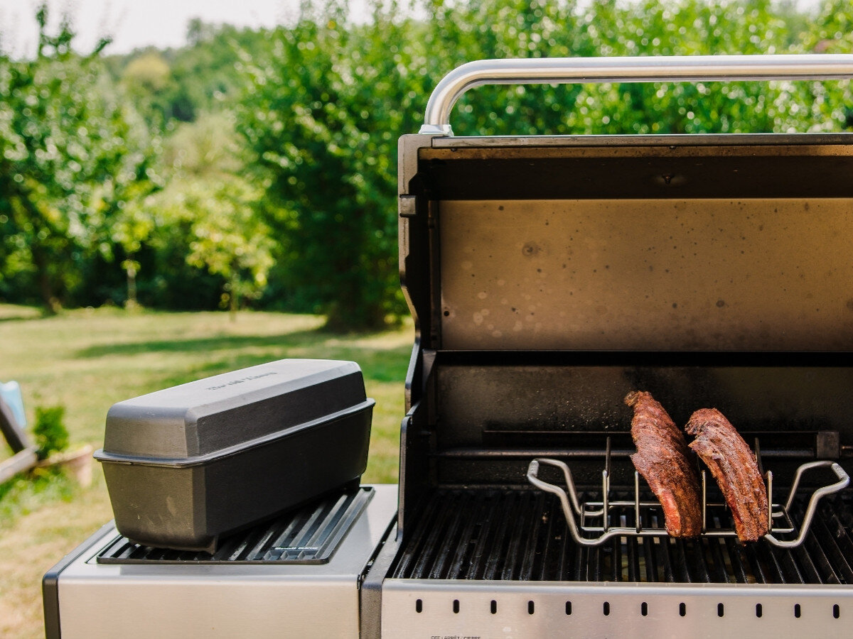 Na zdjęciu widoczny jest grill z otwartą pokrywą, na którym znajdują się dwa kawałki mięsa w stojaku. Obok grilla stoi zamknięty pojemnik na bocznej półce.