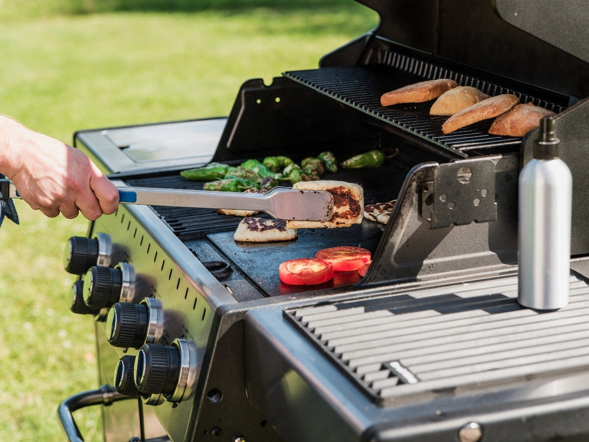 Osoba używa szczypiec do obracania jedzenia na grillu, na którym znajdują się warzywa i pieczywo. Obok grilla stoi metalowa butelka.