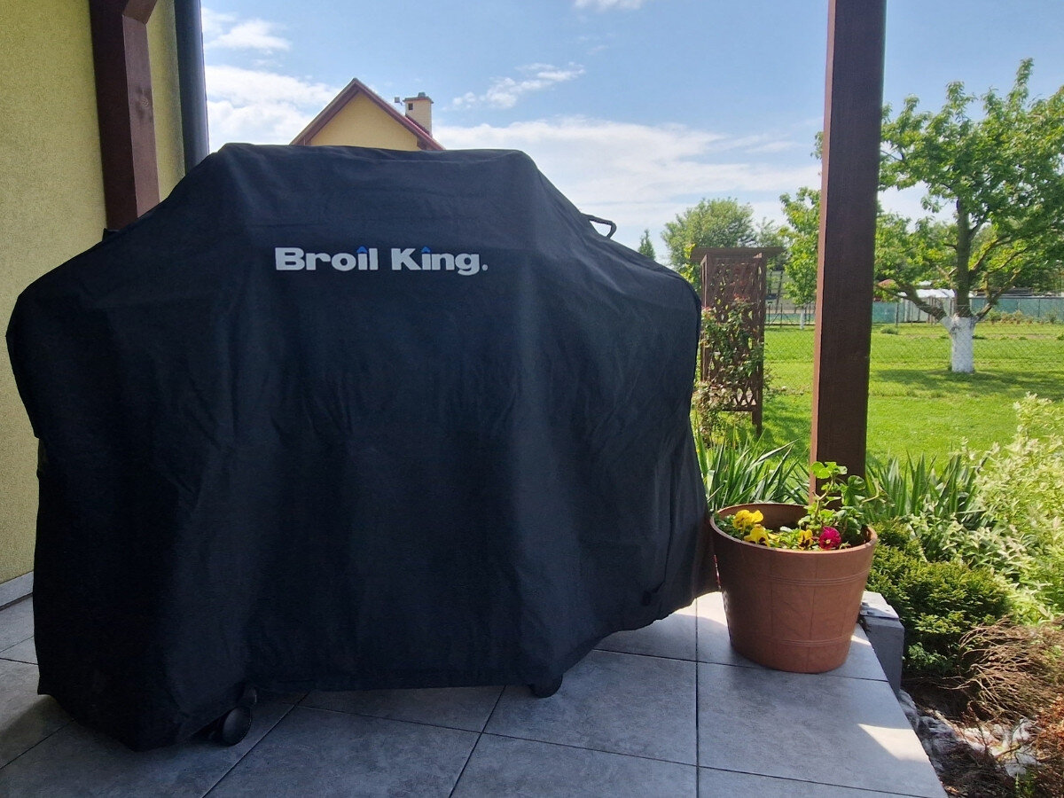 Na tarasie stoi grill przykryty czarnym pokrowcem z widocznym napisem \'Broil King\'. Obok znajduje się donica z kwiatami, a w tle widać ogród z drzewami.