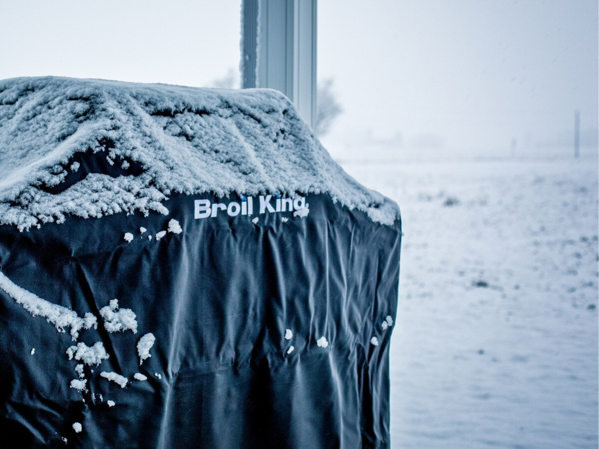 Czarny pokrowiec na grilla pokryty śniegiem z widocznym napisem \'Broil King\'. W tle za oknem widać zaśnieżony krajobraz.