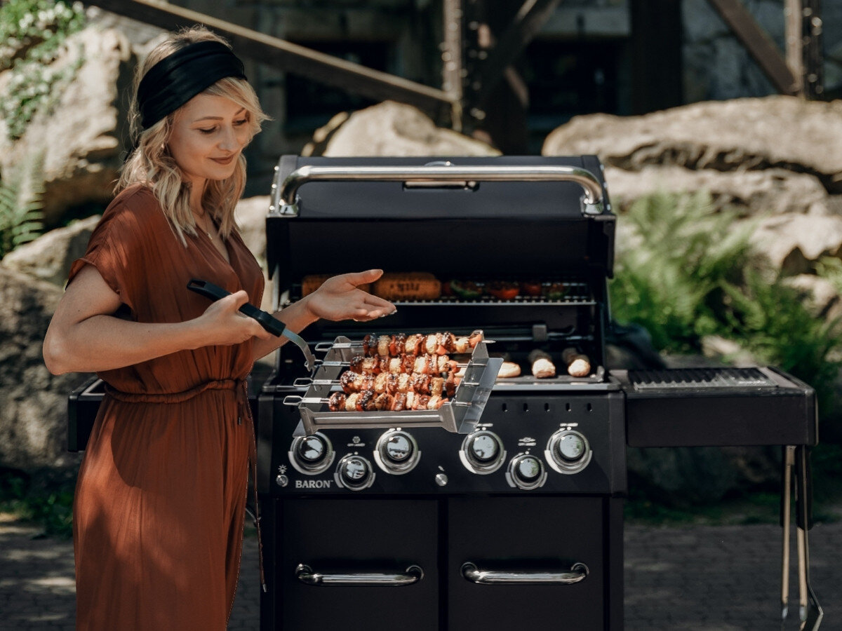 Osoba stoi przy dużym grillu, trzymając zestaw do szaszłyków z kilkoma szaszłykami. Grill jest otwarty, a na ruszcie widać różne potrawy.