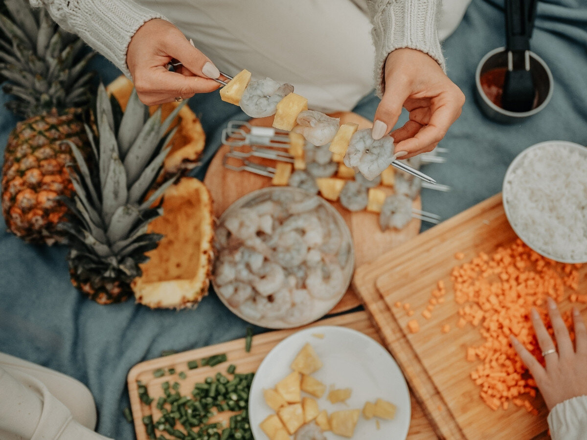 Osoba trzyma szpikulec z krewetkami i kawałkami ananasa nad deską do krojenia z różnymi składnikami. W tle widać ananasa, miski z krewetkami i ryżem oraz deskę z pokrojoną marchewką i szczypiorkiem.