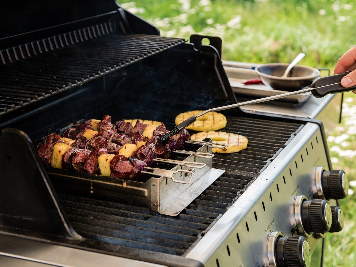 Na grillu znajduje się zestaw do szaszłyków z kawałkami mięsa i ananasa. Obok widoczne są pokrętła do regulacji temperatury oraz miska z łyżką.