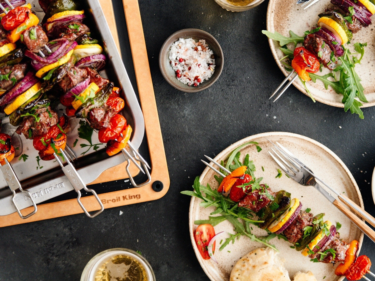 Na stole znajduje się zestaw do szaszłyków z metalowymi szpikulcami, na których umieszczone są kawałki mięsa, papryki, cebuli i cukinii. Obok leżą talerze z gotowymi szaszłykami, rukolą i kawałkami pomidora, a także miseczka z solą i pieprzem.