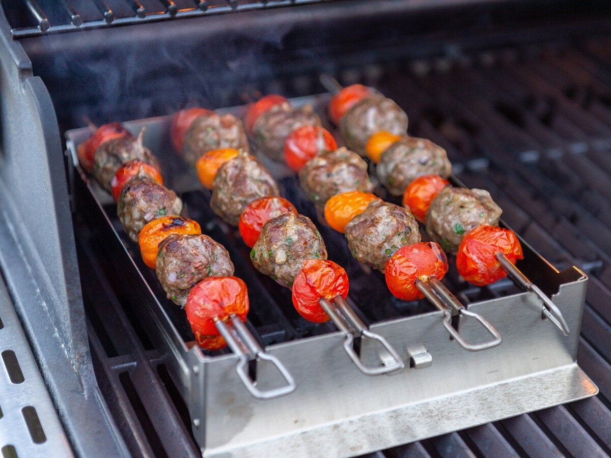 Na grillu znajduje się zestaw do szaszłyków z metalowymi szpikulcami, na których umieszczone są kawałki mięsa i pomidory. Szaszłyki są umieszczone w metalowym stojaku, a z grilla unosi się lekki dym.