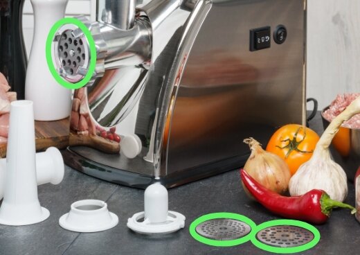 Maszynka do mielenia mięsa MMM-07M z wymiennymi sitkami o różnych oczkach, akcesoria i świeże warzywa na blacie kuchennym.