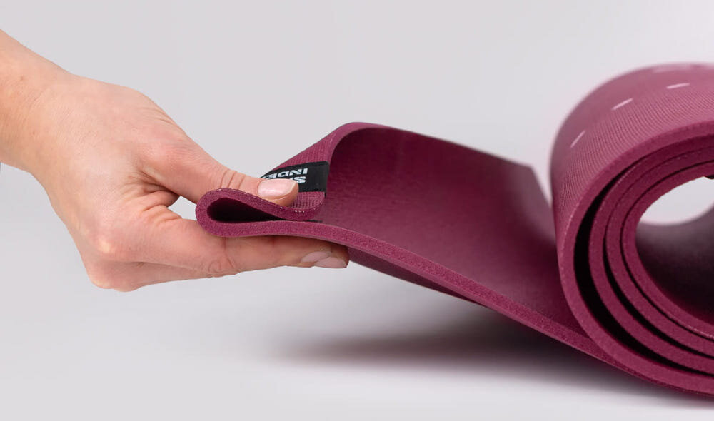 Mata do jogi TIGUAR Yoga Basis Fioletowy jasne tło zwinięta mata osoba trzyma jeden koniec maty w palcach widok z boku wymiary 183 x 60 x 0,5 cm nie ślizga się po podłożu pewność ruchów stabilne oparcie dłoni i stóp zwiększa bezpieczeństwo płynniejszy trening i mniejsze ryzyko niekontrolowanych poślizgów