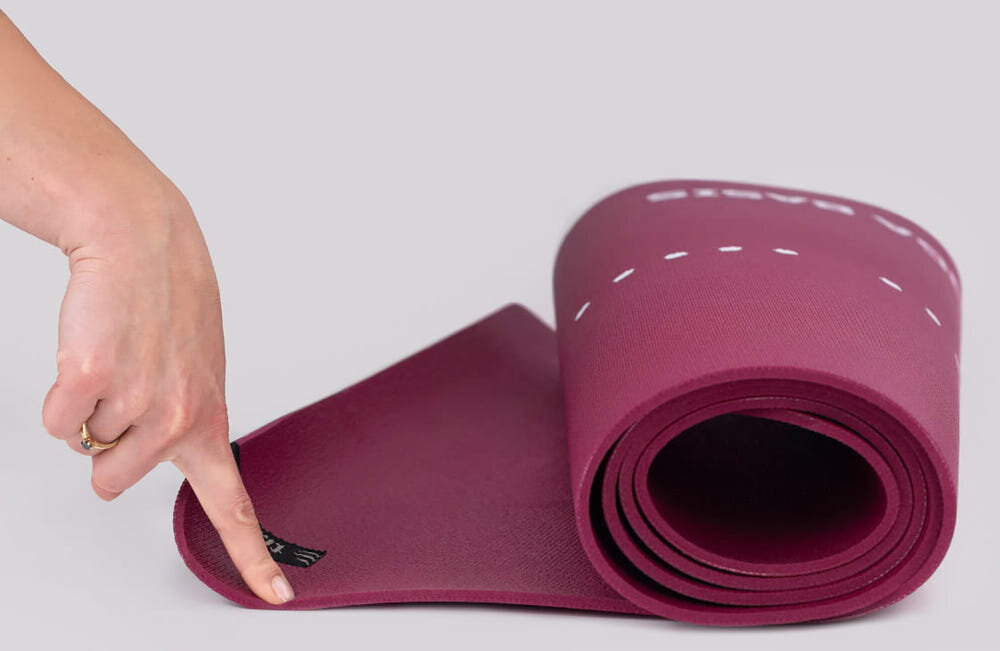 Mata do jogi TIGUAR Yoga Basis Fioletowy jasne tło zwinięta mata osoba trzyma jednym palcem jeden koniec maty widok z boku regularne przecieranie maty wilgotną ściereczką z wodą lub delikatnym środkiem czyszczącym poprawia przyczepność powierzchni każdy kolejny trening odbywa się w bardziej higienicznych warunkach