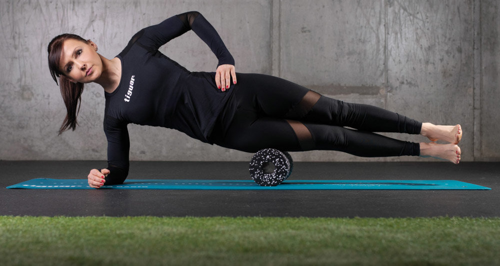 Mata do jogi TIGUAR Yoga Basis Niebieski sala ćwiczeń ciemna podłoga osoba leży na macie biodra ma na rolerze podparta na jednym ramieniu w tle ciemnoszara ściana do wykonywania ćwiczeń jogi stretchingu mobilizacji stabilne i bezpieczne podparcie na różnych typach podłoża odpowiednie kontrola ciała podczas treningu amortyzacja zmniejsza nacisk