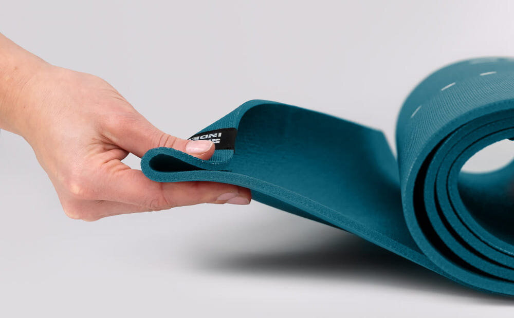 Mata do jogi TIGUAR Yoga Basis Niebieski jasne tło zwinięta mata osoba trzyma jeden koniec maty w palcach widok z boku wymiary 183 x 60 x 0,5 cm nie ślizga się po podłożu pewność ruchów stabilne oparcie dłoni i stóp zwiększa bezpieczeństwo płynniejszy trening i mniejsze ryzyko niekontrolowanych poślizgów