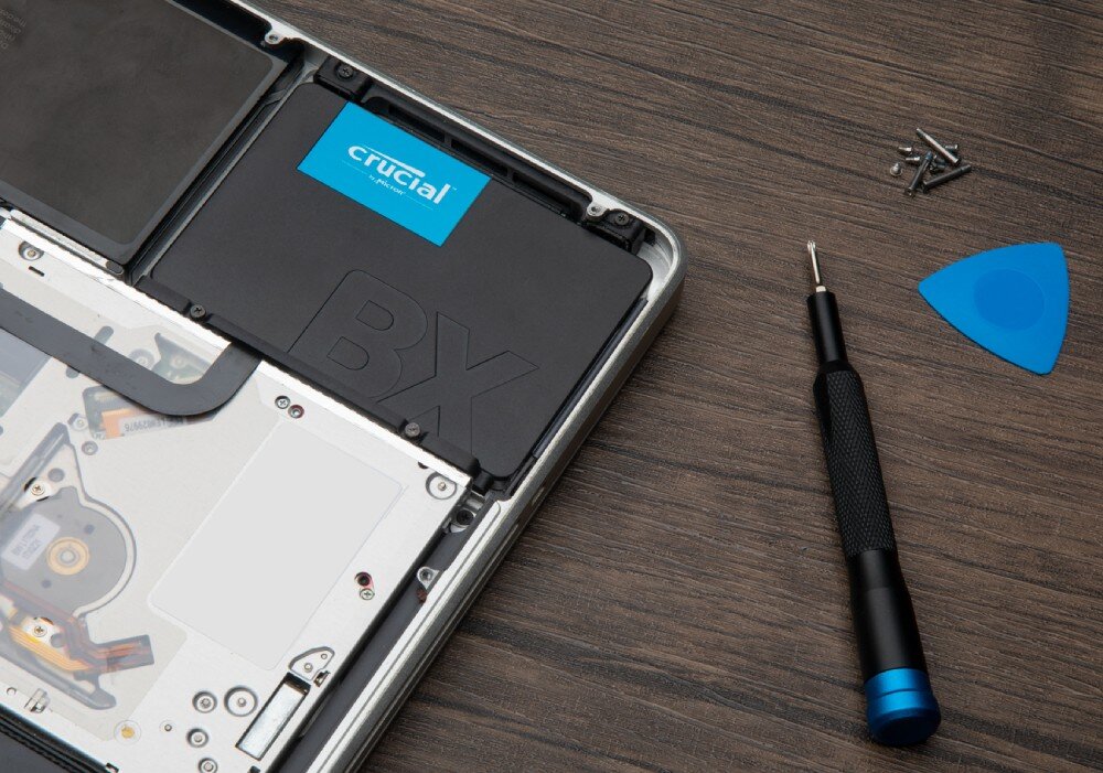 Dysk CRUCIAL BX500 Czarny dysk SSD zamontowany w laptopie, obok śrubokręt, śruby i plastikowe narzędzie na drewnianym blacie, interfejs, podłączenie, kompatybilność