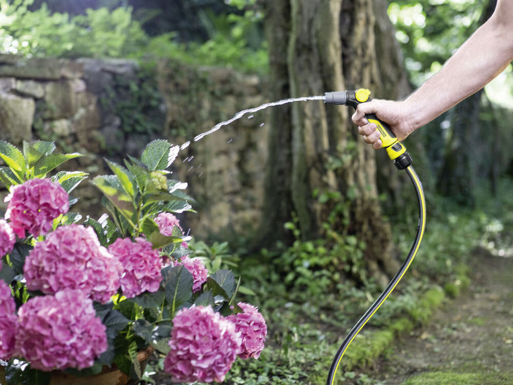 Wąż ogrodowy KARCHER PrimoFlex Plus 1/2 50 m 2.645-319.0, podlewanie, systemy, zasilanie, odporność, konstrukcja, w tle mur i ogród, na pierwszym planie osoba podlewa kwiat w doniczce
