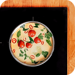 Na obrazie widać patelnię z gotującą się potrawą, w której znajdują się pomidory, plasterki chili, liście ziół i plaster cytryny. Patelnię umieszczono na czarnej płycie grzewczej, a wokół niej widoczny jest drewniany blat.