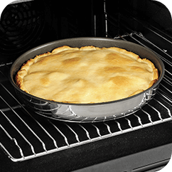 Okrągła patelnia z ciastem w środku znajduje się w piekarniku na metalowej kratce. Ciasto ma złocistą, lekko wypukłą powierzchnię.