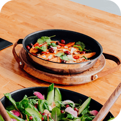 Na drewnianym stole stoi patelnia z jedzeniem, obok znajduje się miska z sałatką. Patelnię otacza drewniana podstawka.