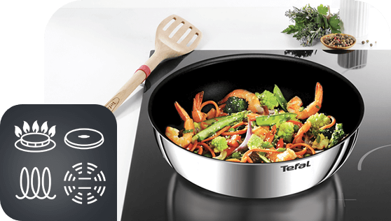 Na obrazie widoczna jest patelnia z logo Tefal, na której znajdują się warzywa i krewetki. Obok patelni leży drewniana łopatka, a w lewym dolnym rogu znajduje się ikona przedstawiająca różne rodzaje kuchenek.