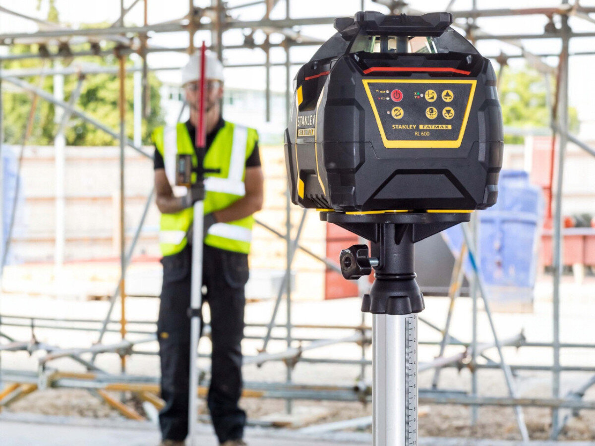 Niwelator laserowy STANLEY FMHT77449-1, laser, budowa, pomiar, poziom, Niwelator Stanley Fatmax RL 600L na statywie, w tle pracownik w kamizelce odblaskowej.
