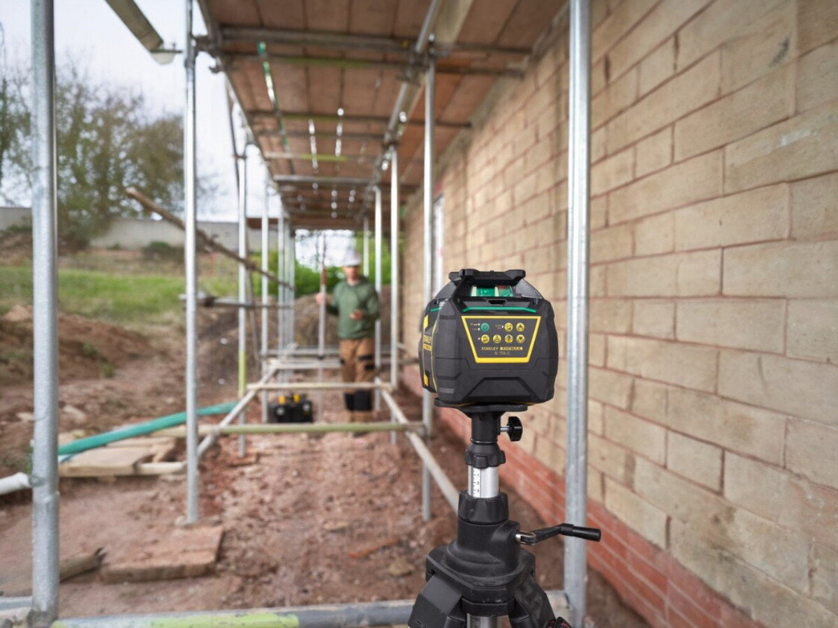 Niwelator laserowy STANLEY FMHT77448-1, bateria, 30godzin, ostrzeżenie, statyw, Pracownik z łatą mierniczą na budowie, niwelator na pierwszym planie.