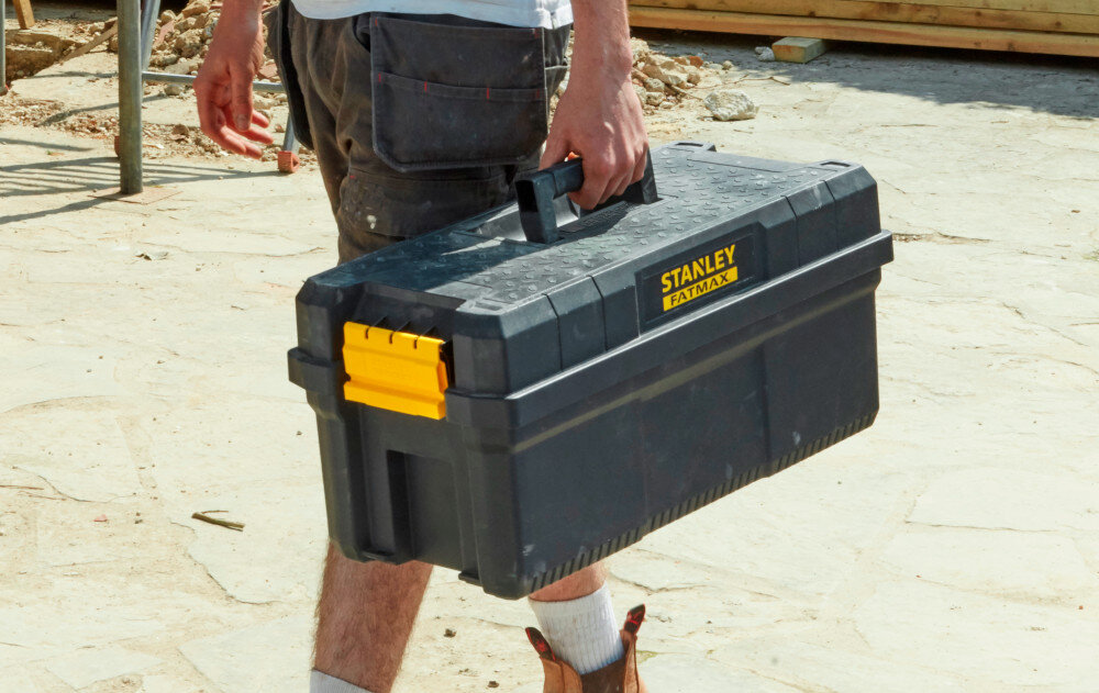Skrzynka narzędziowa STANLEY 81083-1 plac budowy osoba niesie skrzynię w dłoni wymiary 64 × 29,6 × 28,7 cm waga 6,2 kg ergonomiczny uchwyt