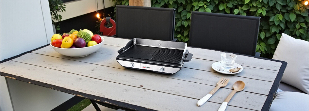 Grill elektryczny GASTROBACK 42523, przeciwrozpryskowa, otoczenie, szpatuła, potrawy, grill stoi na stole tarasowym w tle owoce i sztuće