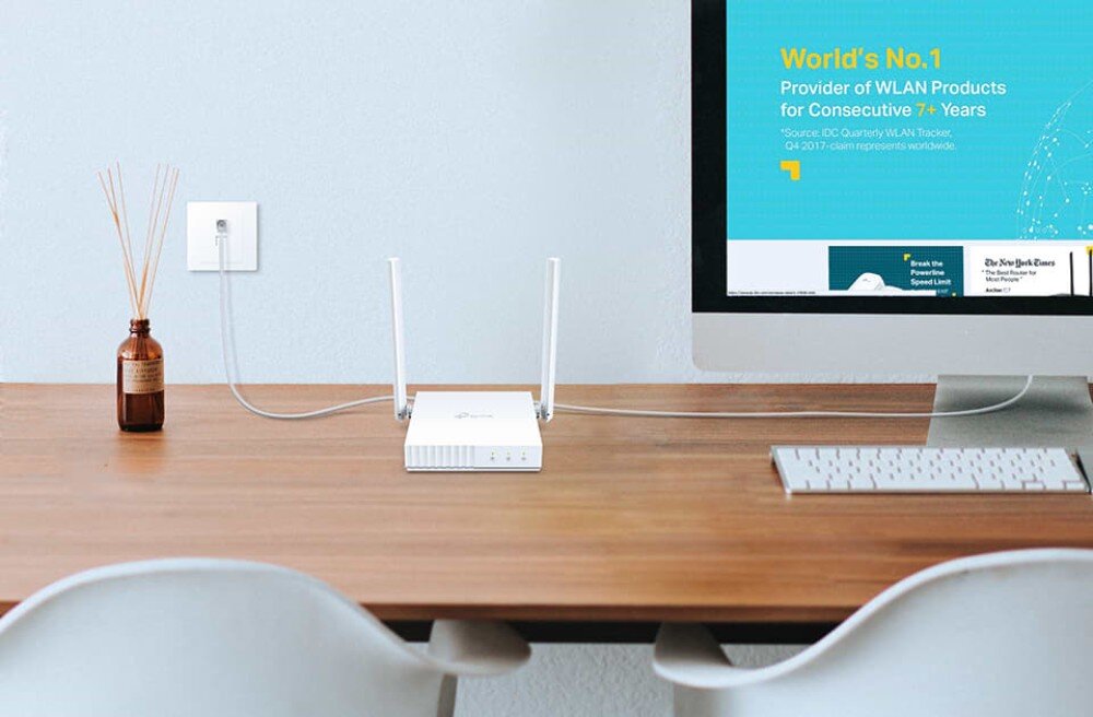 Router TP-LINK TL-WR844N Router ustawiony na drewnianym biurku obok monitora, klawiatury i dekoracyjnej buteleczki z patyczkami zapachowymi, tryby działania