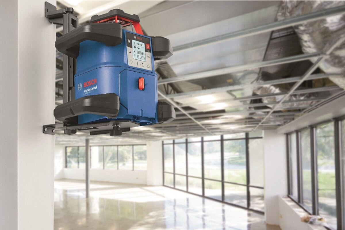 Na obrazie widać niebieski laser obrotowy zamontowany na ścianie w dużym, jasnym pomieszczeniu z oknami. Na urządzeniu widoczne jest logo \'BOSCH Professional\'.