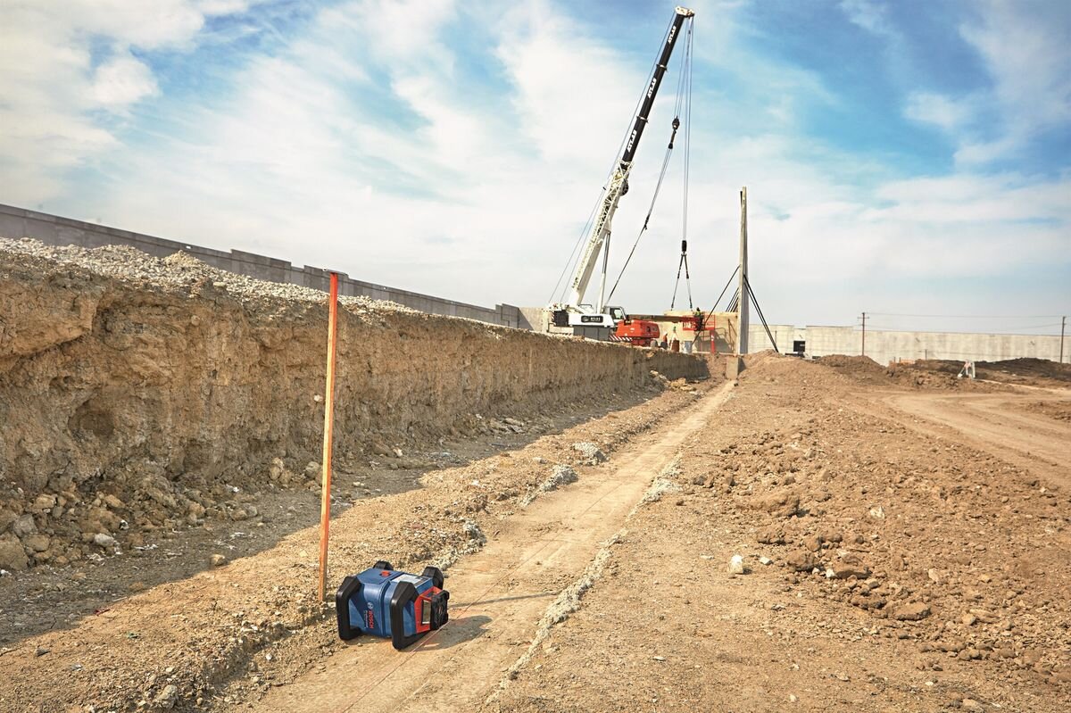 Na placu budowy widać niebieskie urządzenie z czerwonymi elementami, obok którego stoi pomarańczowy pręt. W tle znajduje się dźwig oraz betonowy mur.
