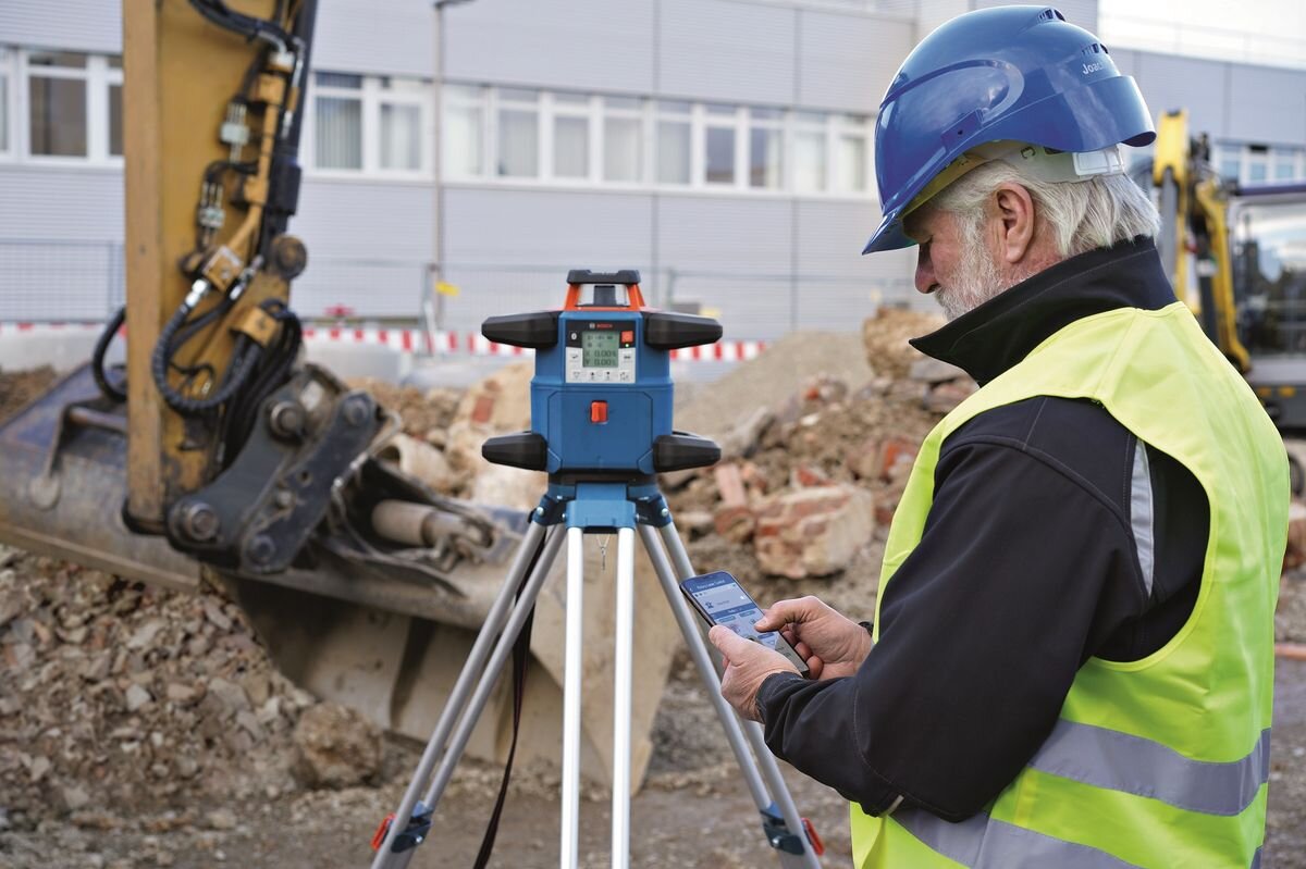 Osoba w kasku i kamizelce odblaskowej obsługuje urządzenie laserowe na statywie na placu budowy. W tle widoczne są maszyny budowlane i budynek.