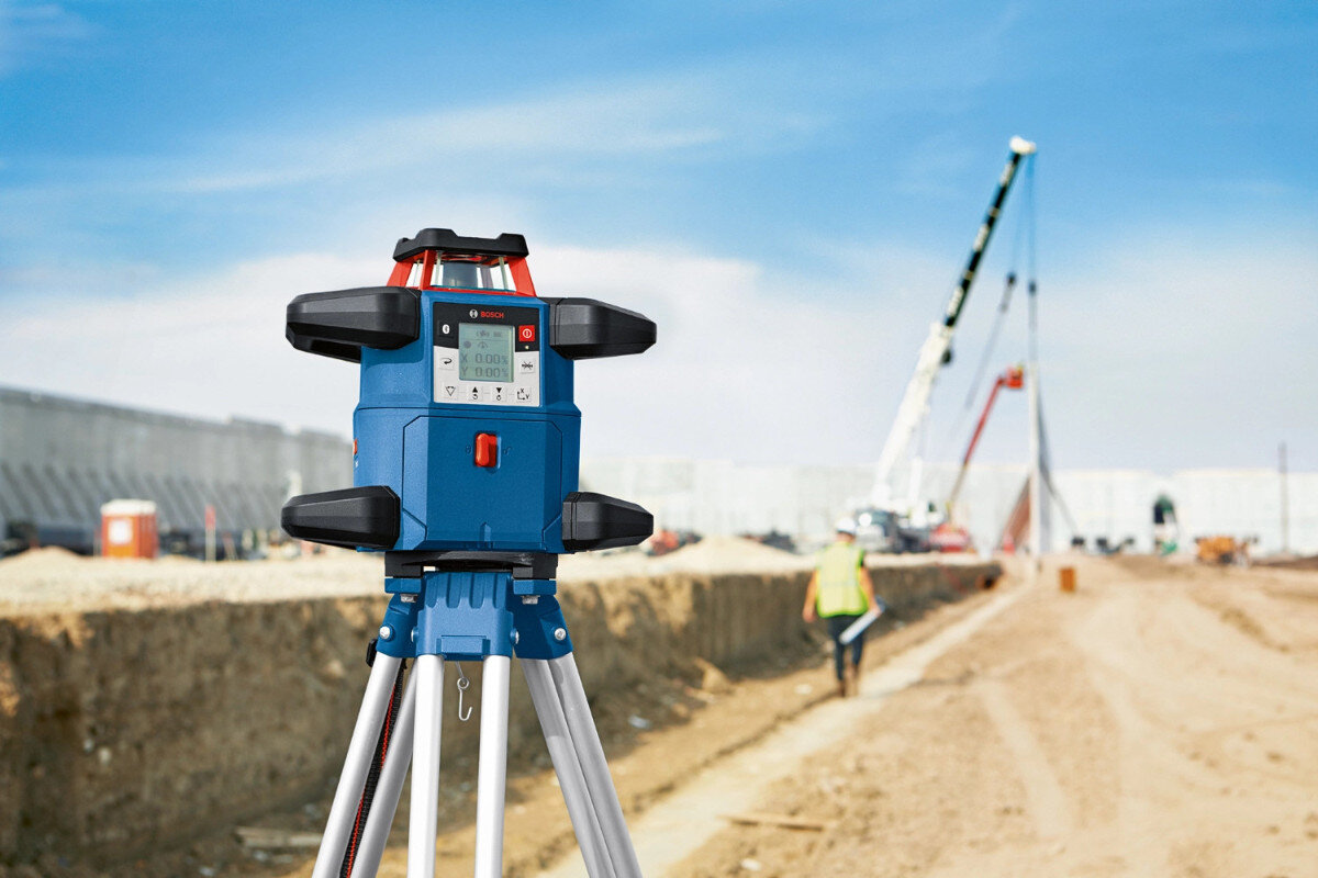 Na placu budowy znajduje się laser obrotowy na statywie z widocznym logo Bosch. W tle widać pracownika i dźwig.