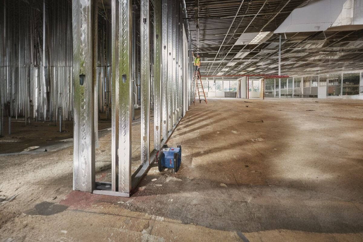 Wnętrze budynku w trakcie budowy z widocznymi metalowymi stelażami i urządzeniem na podłodze. W tle osoba na drabinie pracuje przy suficie.