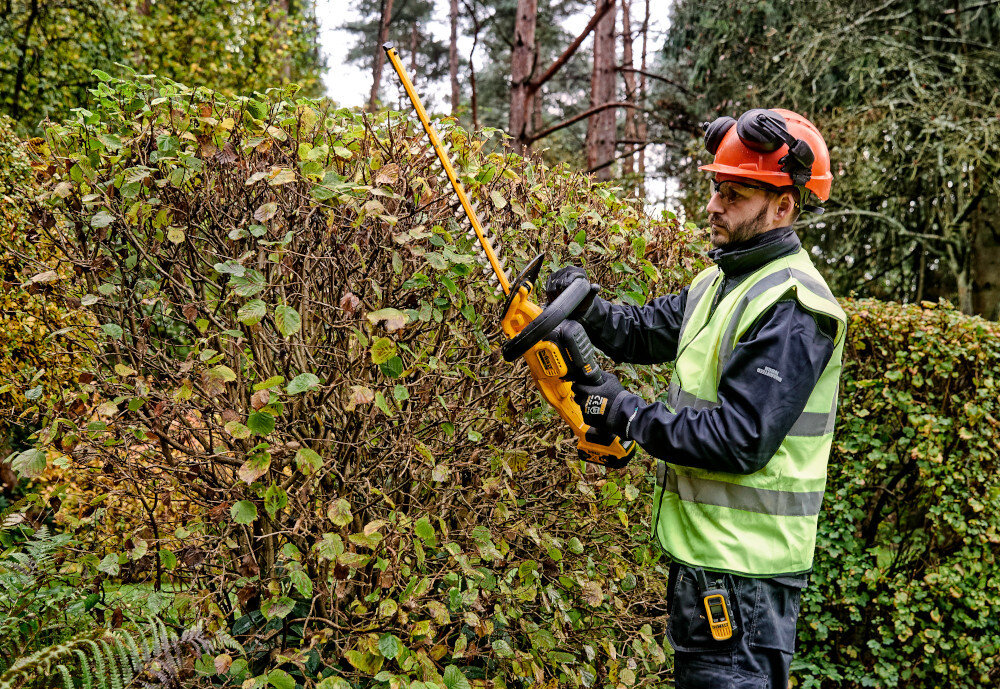 Nożyce do żywopłotu DEWALT DCMHT563P1 18V akumulatorowe ogród osoba przycina żywopłot w tle drzewa niebo waga około 2,9 kg bez akumulatora ergonomiczne uchwyty pokryte gumą łatwe prowadzenie narzędzia ogranicza obciążenie ramion pewny chwyt kontrola nad ruchem ostrza