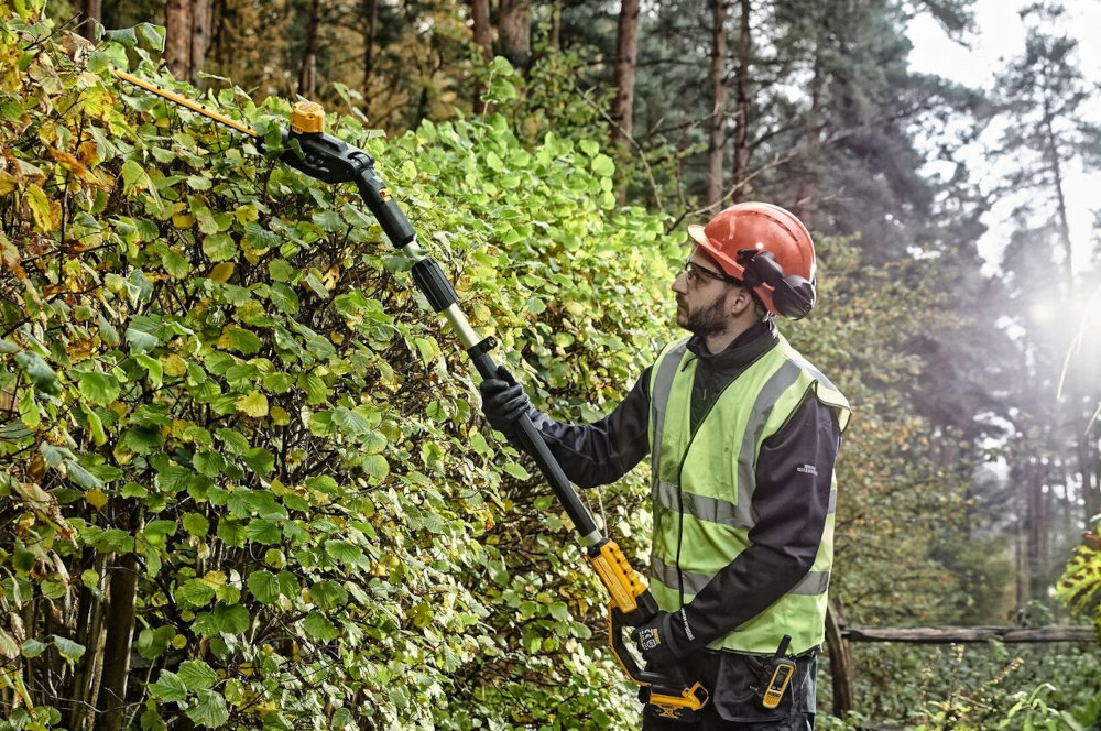 Nożyce do żywopłotu DEWALT DCMPH566P1-QW 550mm 18V akumulatorowe ogród osoba stoi na trawniku tnie żywopłot w tle drzewa krzewy do 75 minut na jednym ładowaniu akumulatora ułatwia utrzymanie równego rytmu cięcia wygoda użytkowania czas ładowania wynosi około 75 minut