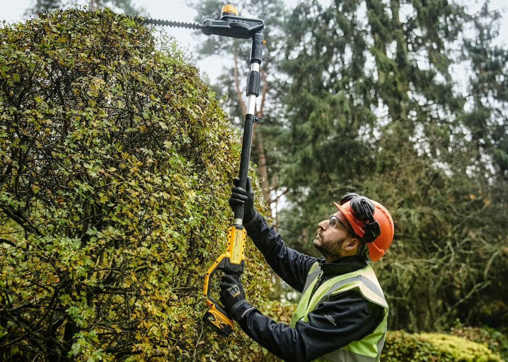 Nożyce do żywopłotu DEWALT DCMPH566P1-QW 550mm 18V akumulatorowe ogród osoba stoi na trawniku tnie żywopłot w tle drzewa krzewy wysięgnik o długości 2,15 m maksymalny zasięg roboczy do 3,35 m przycinanie wysokich żywopłotów i głębokich krzewów bez zmiany stanowiska pracy