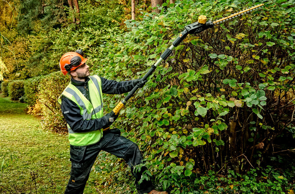 Nożyce do żywopłotu DEWALT DCMPH566P1-QW 550mm 18V akumulatorowe ogród osoba stoi na trawniku tnie żywopłot w tle drzewa krzewy pasek na ramię równomiernie rozłożony ciężar nożyc zmniejsza obciążenie ramion i pleców większa precyzja cięcia i komfort wspiera ergonomię pracy ułatwia manewrowanie poprawia wydajność