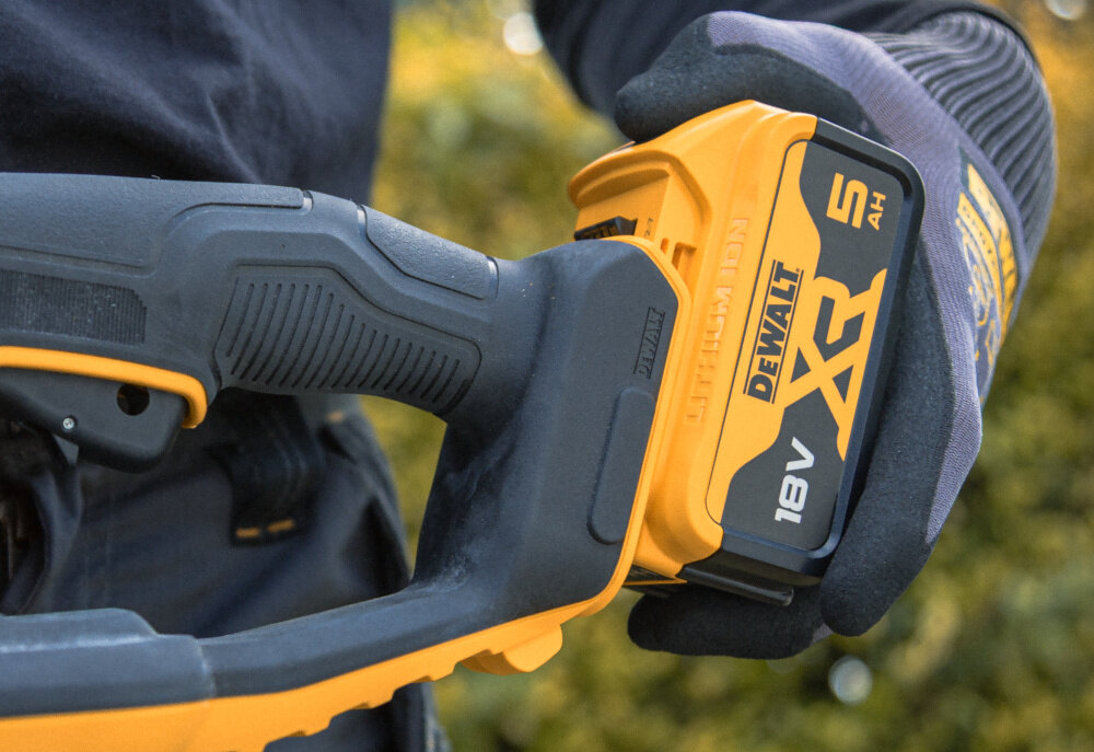 Nożyce do żywopłotu DEWALT DCMHT563N-XJ 18V akumulatorowe ogród osoba trzyma w dłoniach nożyce wkłada akumulator na miejsce należą do systemu DEWALT 18 V XR® wygoda pracy bez przewodu parametry zbliżone do narzędzi sieciowych oszczędność czasu i pieniędzy jeden akumulator można wykorzystać w innych narzędziach