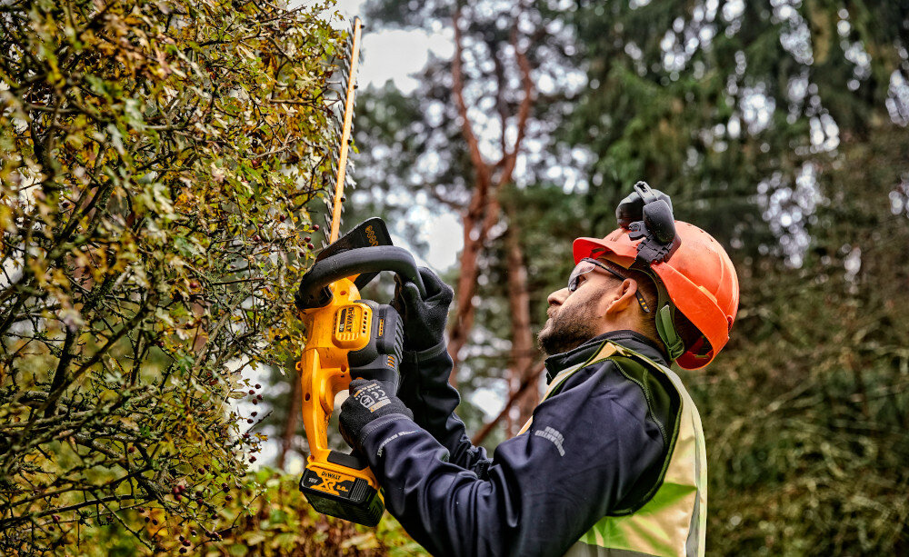 Nożyce do żywopłotu DEWALT DCMHT563N-XJ 18V akumulatorowe ogród osoba przycina żywopłot w tle drzewa niebo do 75 minut na jednym ładowaniu akumulatora o pojemności 5,0 Ah ciągłość pracy ma realny wpływ na tempo realizacji wygoda użytkowania