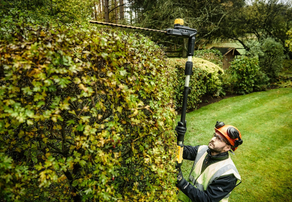 Nożyce do żywopłotu DEWALT DCMPH566N-XJ 550mm 18V akumulatorowe ogród osoba stoi na trawniku tnie żywopłot w tle drzewa krzewy wygodne cięcie pielęgnacja wysokiej zieleni bezpieczeństwo wygoda